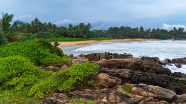 Sri Lanka 'nın sakin kıyı manzarası bulutlu. Bereketli yeşillik, bulutlu bir günde Sri Lanka 'da uzak bir plajın kıyısına vuran yumuşak dalgalarla kayalık kıyı şeridiyle buluşur..