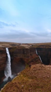 İnsanlar tehlikeli kayanın kenarında durup güzel şelaleyi seyrediyorlar. Beyaz su akışının havadan perspektifi zemine çarpıyor ve yukarı doğru beyaz buhar yaratıyor. Dikey video.