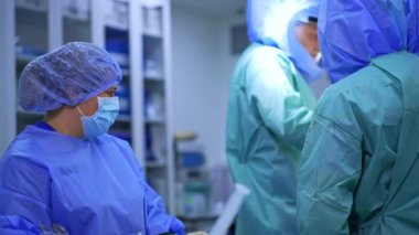 Koruyucu kasklı bir erkek cerrah, süngerle kana bulanmış bir forsepsi geçer. Ameliyat hemşiresi temiz bir sünger verdi. Bornozunda kan lekeleri olan tanınmamış bir doktor sırtı kameraya dönük duruyor..