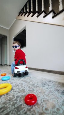 Güneş gözlüğü takan sevimli çocuk oyuncak araba kullanıyor. Çocuk oyuncak piramit halkalarının arasında geziyor. Düşük açı. Dikey video.