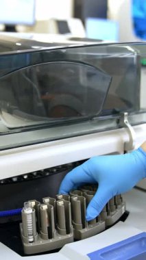 Tıbbi laboratuvar asistanı test tüpüyle makineye bir stand yerleştiriyor. Eldivenli kadın, test prosedürünü başlatmak için ekipmanın kapağını kapatıyor. Dikey video