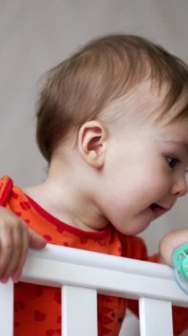 Güzel beyaz erkek bebek beyaz karyolada kalıyor. Tatlı çocuk emzik ile oynuyor yere atıyor ve düşüşünü seyrediyor. Düşük açı. Dikey video