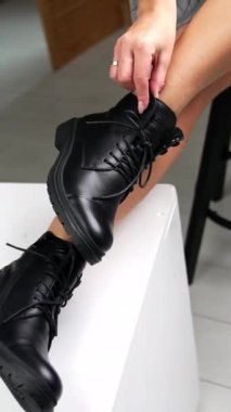 Ayaklarını beyaz küpün üzerinde tutan bir taburede oturan kadın. Modern fotoğraf stüdyosunda bağcıklı siyah çizmeleri gösteren bir model. Dikey video