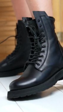 Kadın ayaklarında bağcıkları olan şık asker botlarının gösterimi. Kameranın önünde şık ayakkabılar giyen bir kadın. Kapatın. Arkaplanda tahta perde var. Dikey video