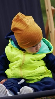 Beyaz çocuk sıcak kıyafetler giyip bahçe sandalyesinde oturuyordu. Bebek dikkatlice etrafına bakıyor, sakince oturuyor. Kapatın. Bulanık doğa arkaplanı. Dikey video