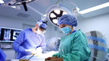 Pozitif kadın hemşire cerrahi standında duruyor ve aletleri ayarlıyor. Cerrahi lensler takan erkek cerrah hastayı ameliyat etmek için parlak lambanın altında duruyor..