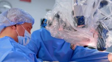 Profesyonel erkek cerrah alet alır. Beyin cerrahı mikroskop ameliyatına bakıyor. Yan görüş. Asistan, şırıngadaki ilaçları operasyon yerine püskürtür..