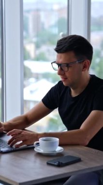 Laptop 'ta çalışan üretken genç bir adam. Erkek serbest çalışan dikkatle ekrana bakıyor ve bir şeyler yazıyor. Kapatın. Dikey video