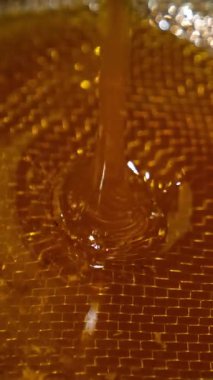 Makineden sızan altın bal akışı. Balmumu parçalarını filtrelemek için metal eleğe bal dökülüyor. Kapatın. Dikey video