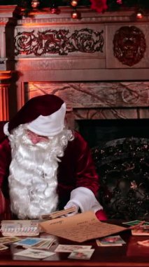 Noel Baba, Noel kartlarını kırmızı tüyle yazmayı bitirir. Noel Baba ayağa kalkar ve güzel süslenmiş bir şöminenin yanına gelir. Dikey video