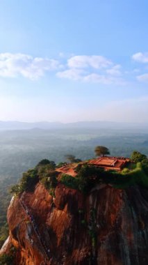 Tepesine merdivenler olan kahverengi dağ ve tepesinde yemyeşil bir yer. Sigirya Kalesi 'nin havadan görünüşü harabe. Arka planda puslu bir manzara. Dikey video.