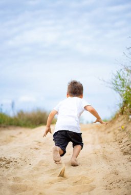 Çocuk güneşli bir yolda koşuyor. Genç bir çocuk neşeli bir şekilde kumlu bir patika boyunca çıplak ayakla koşar. Parlak bir gökyüzünün altında yeşilliklerle çevrilidir..