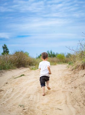 Çocuk kumlu yolda yalınayak koşuyor. Genç bir çocuk mavi gökyüzünün altında çimlerle ve ağaçlarla çevrili kumlu bir yolda çıplak ayakla koşuyor, doğanın güzelliğinin tadını çıkarıyor..