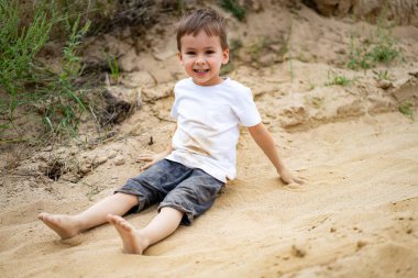 Genç çocuk açık kumda mutlu bir şekilde oynuyor. Kumsalda güneşli bir günün tadını çıkarırken kumsalda oturan bir çocuk, eğlencenin tadını çıkarıyor..
