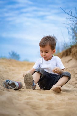 Güneşli bir günde kumda oynayan küçük bir çocuk. Bir çocuk kumlu bir yolda oturuyor, ayakkabısını giymeye odaklanmış açık havada güneşli bir günün tadını çıkarıyor..