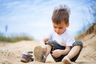 Genç çocuk güneşli bir günde kumla oynuyor. Aydınlık, güneşli bir günde kumlu bir yolda oturan ve yanında ayakkabıları olan bir çocuk kumla ilgilenir..