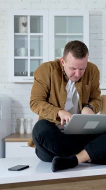 Mutfak masasında oturan ve dizüstü bilgisayarda yazan komik adam. Genç adam uzun süre dizüstü bilgisayarda çalıştı. Evde çalışan bir serbest çalışanın yorgun yüzü. Uzaktan kumanda. Dikey video