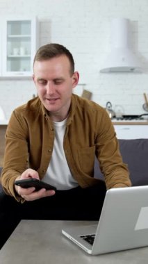 Evde çalışan serbest çalışan bir erkek. Genç adam bir dizüstü bilgisayarın önünde kanepede oturuyor ve mutfak arka planındaki cep telefonuna bakıyor. Dikey video