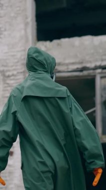 Terk edilmiş bir yerde şaşkın bir kurtulan. Güvenlik üniformalı ve gaz maskeli bir insan yıkılmış binaların arka planına bakıyor. Dikey video