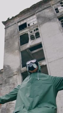 Gaz maskeli adam mahvolmuş bir yerde. Koruyucu giysili ve solunum cihazlı bir insan yıkılmış binanın arka planında kollarını açmış dışarıda oturuyor. Radyoaktif bölge. Dikey video