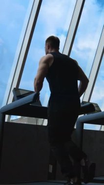 Gökyüzü arka planında koşu bandında bir sporcu. Spor salonundaki spor salonundaki adam simülatörde koşuyor ve pencereden bakıyor. Dikiz aynası. Dikey video