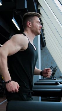 Sporcu koşu bandında koşuyor. Spor salonunun arka planında simülatör üzerinde egzersiz yapan genç vücut geliştirici. Profil görünümü. Dikey video