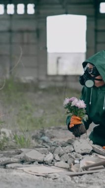 Terk edilmiş bir yerde güvenlik üniformalı bir kadın. Koruyucu giysi ve solunum cihazlı bir bilim adamı yıkık dökük binada çiçek taşıyor. Çevre tehlikede. Dikey video