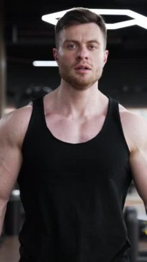 Yakışıklı sporcu spor salonunda yürür. Modern spor salonunda siyah cüppeli genç bir sporcunun portresi. Kaslı vücut geliştirici içeride. Dikey video