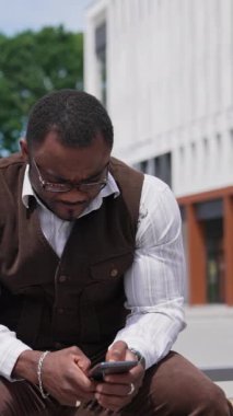 Telefona bakan ciddi siyah derili adam. Takım elbiseli, açık havada oturan ve cep telefonu kullanan Afrikalı Amerikalı adamın portresi. Dikey video