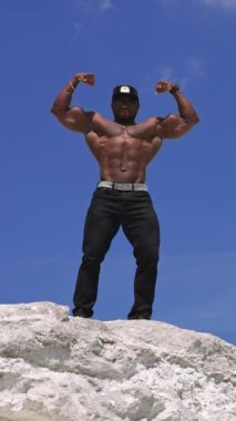 Mavi gökyüzü arka planında poz veren Afrikalı Amerikalı sporcu. Koyu tenli atlet dışarıda güçlü kol kaslarını gösteriyor. Aşağıdan bak. Dikey video