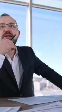 Gülümseyen girişimci ofiste. Masada dizüstü bilgisayarla oturan gözlüklü bir iş adamının portresi. Ofis çalışanı içeride. Dikey video