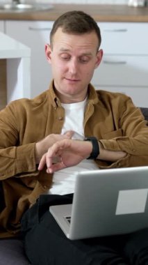 Kanepede yatarken dizüstü bilgisayar kullanan serbest çalışan bir erkek. Genç bir adam evde dizüstü bilgisayarla çalışırken saatine bakıyor ve biriyle konuşuyor. Dikey video