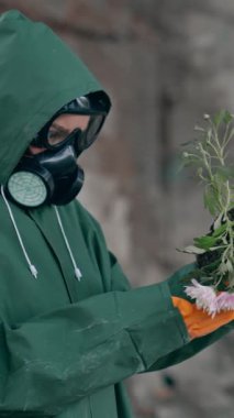 Doğa tehlikede. Güvenlik giysisi ve gaz maskeli bir ekolojist terk edilmiş bir yerde kırık çiçekler tutuyor. Biyokimyasal saldırı. Kirli bir çevre. Dikey video