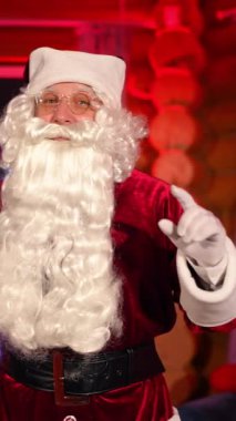 İçeride Noel Baba 'nın portresi. Noel Baba kostümlü, doğal sakallı, yaşlı bir adam Noel 'i kutluyor. Kamerada konuşan yaşlı adam mutlu Noeller diliyor. Dikey video