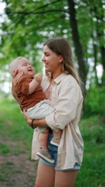 Genç, uzun saçlı bir kadın sevgili bebeğini kucağına alıp sallıyor. Anne ve çocuk doğada zaman geçirirler. Dikey video.