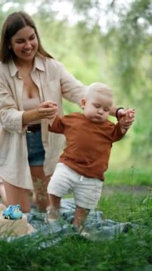 Gülümseyen mutlu beyaz kadın dizlerinin üzerinde oğlunu tutuyor. Küçük bebek ilk adımı atmaya çalışıyor. Dikey video.