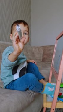 Şirin çocuk elinde keçeli kalemle kanepede oturuyor. Bebek beyaz tahtaya çizerken kalemleri değiştiriyor. Dikey video.