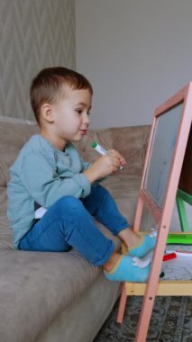 Koyu renk saçlı sevimli çocuk beyaz tahtaya renkli kalemler çiziyor. Evde yaratıcı çocuk çizimi. Dikey video.