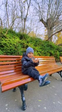 Neşeli gülümseyen bebek tahta bankta oturuyor. Mutlu çocuk el sallayarak oynuyor. Dikey video.