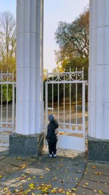 Sıcak ceket ve şapka giyen çocuk kapıyı açtı. Çocuk sonbaharda parkın girişinde oynuyor. Dikey video.