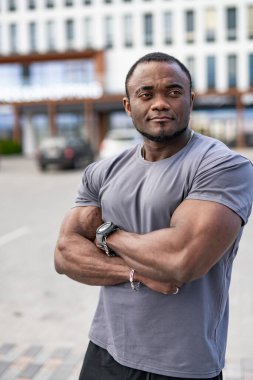 Kendine güveni olan bir adam günlük kıyafetlerle dışarıda poz verir. Kolları çapraz duran kaslı bir adam modern bir açık hava ortamında güç ve kendine güven sergiliyor..