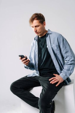 Tepedeki adam akıllı telefonu kontrol ediyor. Bir adam minimalist bir alanda günlük giysiler giyerken akıllı telefonuna odaklanmış bir şekilde bir kaide üzerinde oturuyor..