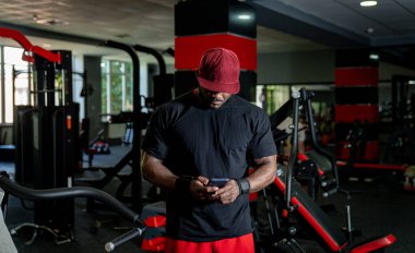 Spor salonundaki adam antrenmandan sonra telefonunu kontrol ediyor. Şapka takan ve spor kıyafeti giyen bir adam modern bir spor salonunda yoğun bir çalışmadan sonra telefonunu kontrol ediyor..