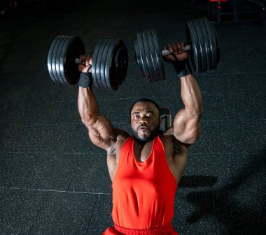 Adam spor salonunda dambıl ütülüyor. Kas yığını bir adam spor salonunda başınızın üstüne ağır dambıllar kaldırır, günün herhangi bir saatinde ağırlık çalışması sırasında güç gösterisi yapar..