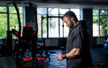 Spor salonunda telefonuna bakan bir adam. Bir adam spor salonunda duruyor, telefonuna odaklanmış ve bir sonraki egzersiz rutinine hazırlanıyor..