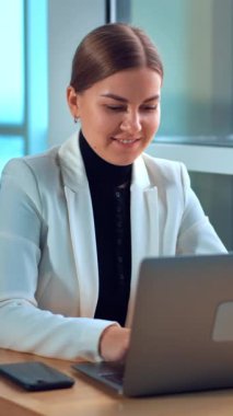 Dizüstü bilgisayarının önünde oturan beyaz ceketli genç bir kadın. Gülümseyen bayan bilgisayarda bir şeyler yazıyor ve düşünceli bir şekilde pencereye bakıyor. Dikey video