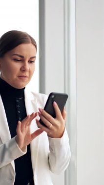 İş kadını pencerede duruyor ve telefon görüşmesi yapıyor. Beyaz ceketli kadın telefonuna bakar, eliyle konuşur ve el kol hareketi yapar. Dikey video