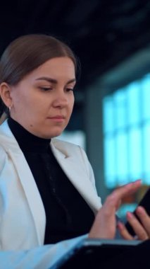 Beyaz ceketli iş kadını rahat koltuğunda oturup telefonuna odaklanıyor. Beyaz bir kadın akıllı telefonunu kullanarak çalışıyor. Kapatın. Arkaplan bulanık. Dikey video