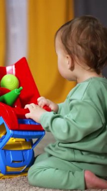 Tatlı çocuk yumuşak halıda parlak bir kamyonla oynuyor. Yürüyen çocuk, oyuncakları arabadan çıkarmak için arabayı çeviriyor. Arkaplan bulanık. Dikey video