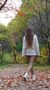 Güzel doğada yavaşça yürüyen uzun saçlı kızı takip ediyorum. Sonbahar parkının yanından geçen beyaz ceketli ve bej tozlu esmer kadın. Dikey video